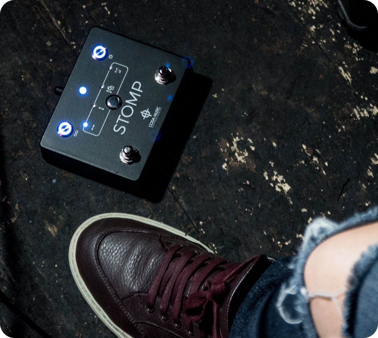 Black stomp box pedal on a dark surface with a person wearing shoes.