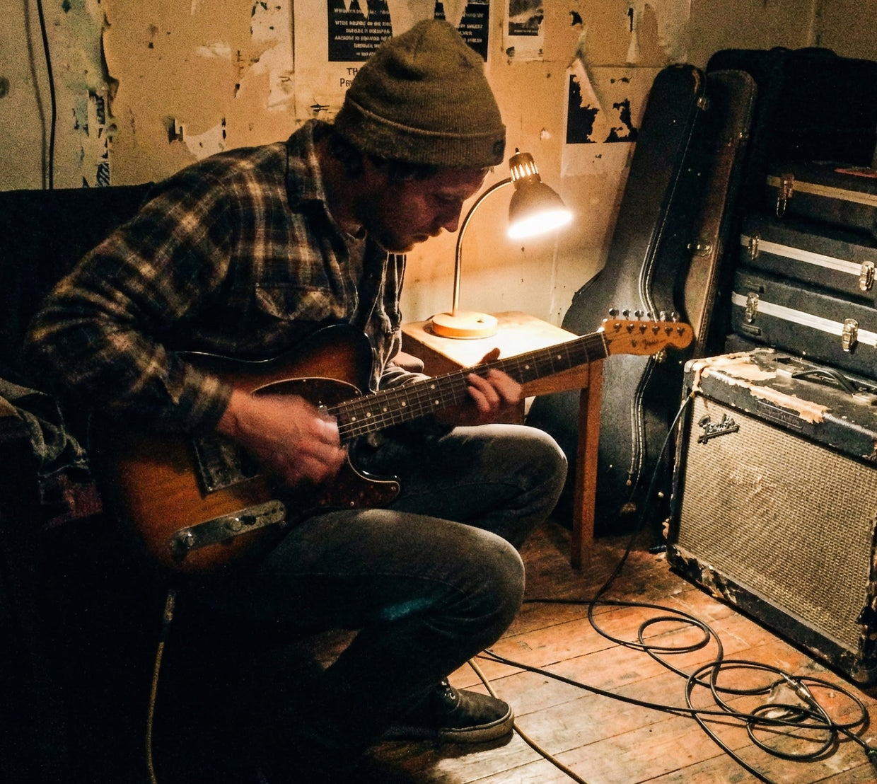 Musician playing an electric guitar beside amplifiers and equipment in a rehearsal room