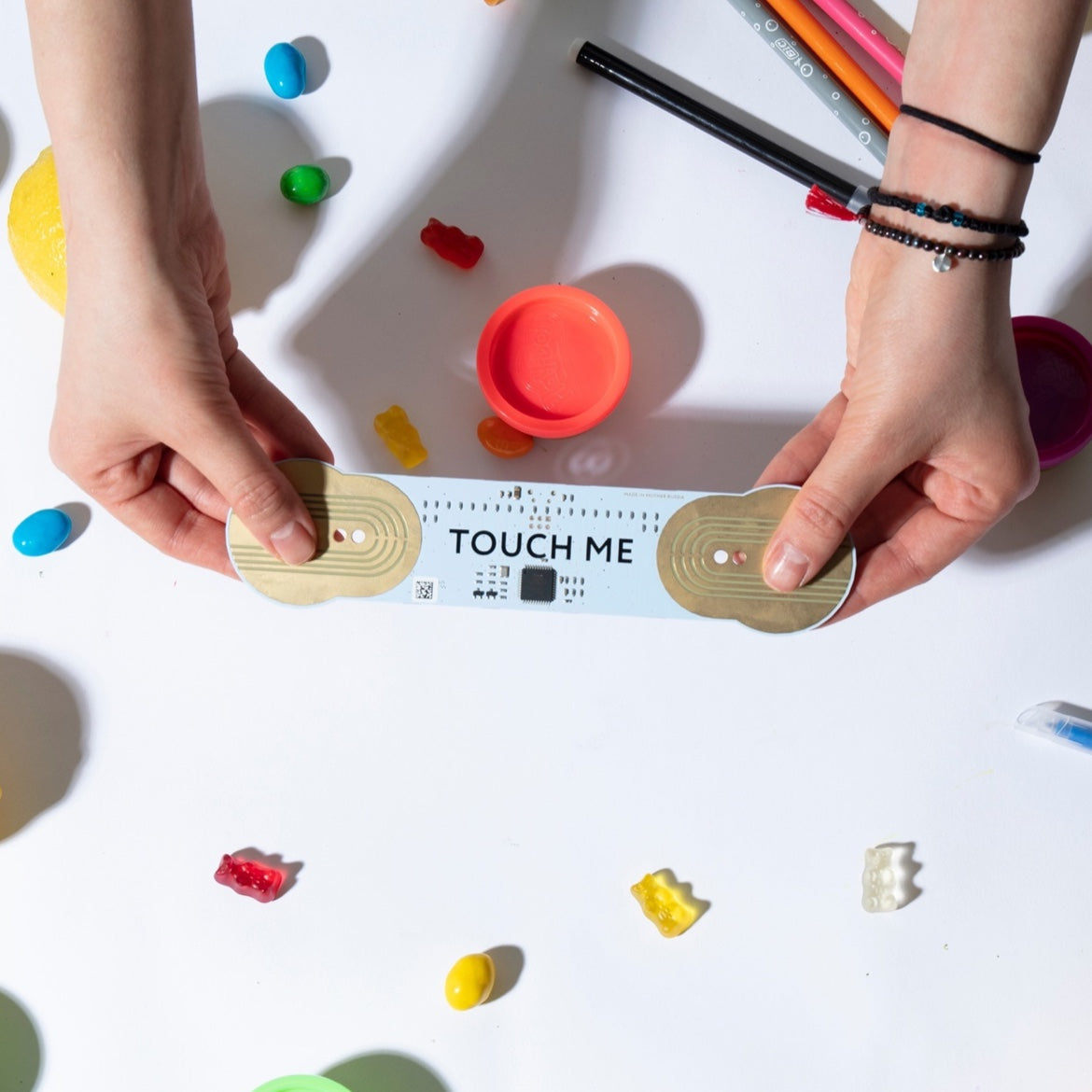Hands holding the TouchMe MIDI instrument over a table with colorful objects